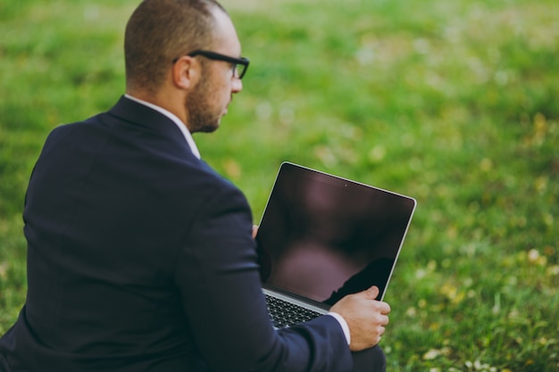 Close up young businessman in suit, glasses. Man sit on soft pouf, working on laptop pc computer in city park on green lawn outdoors on nature. Mobile Office, business concept. Back view. Copy space.