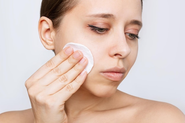 Close up of young caucasian woman cleaning her face with cotton pad isolated on a white background