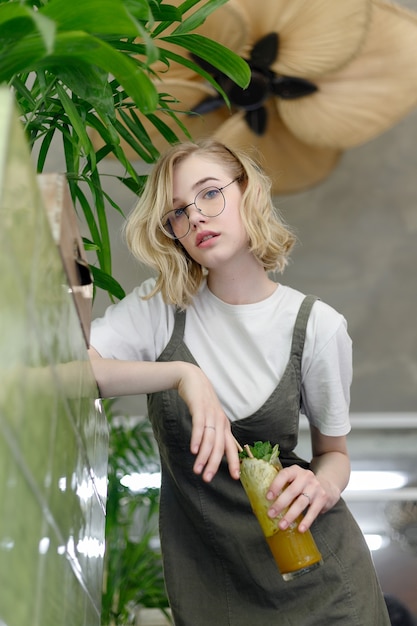 Close up young cheerful woman in dress happily looking in camera with big green leaves on background in cozy green cafe. Girl in glasses. holding a drink in his hands