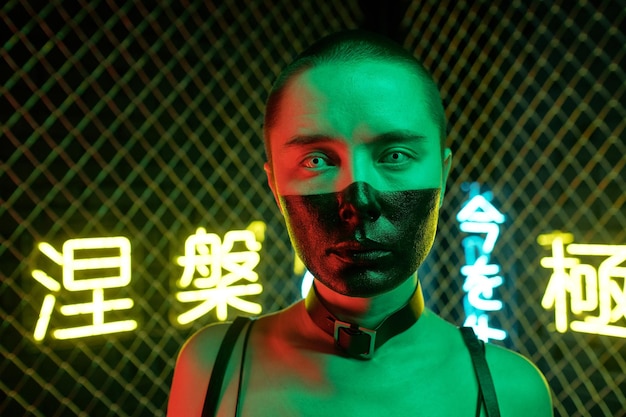 Close-up of young cyberpunk woman with face painted with black color looking at camera against the neon hieroglyphs meaning nirvana and western paradise