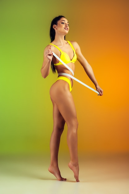 Close up of young fit and sportive woman with measurer in stylish yellow swimwear on gradient wall perfect body ready for summertime