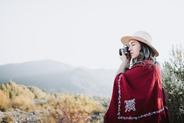 Close up of young latin woman wearing a red poncho and a hat taking photos with a vintage camera