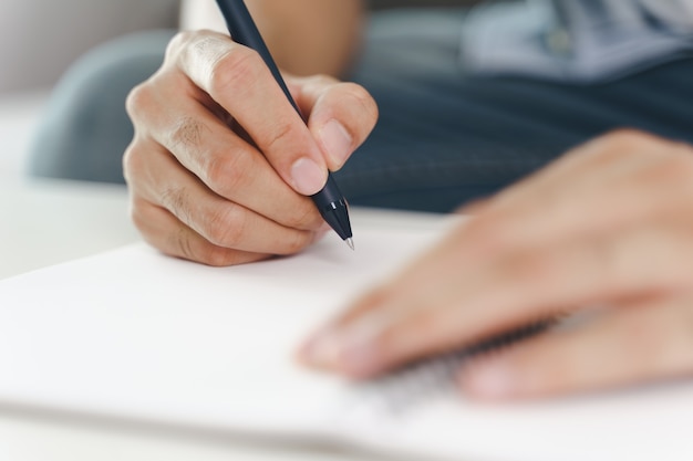 Close up of young man in casual cloth hands writing down on the notepad, notebook using ballpoint pen on the table.