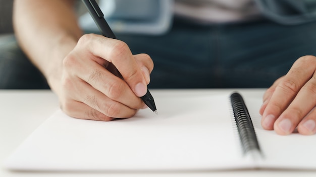 Close up of young man in casual cloth hands writing down on the notepad, notebook using ballpoint pen on the table.