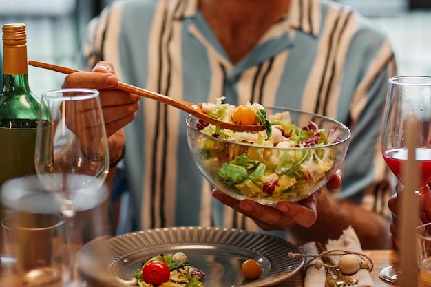 Close up of young man holding salad bowl while enjoying dinner party in cozy setting copy space