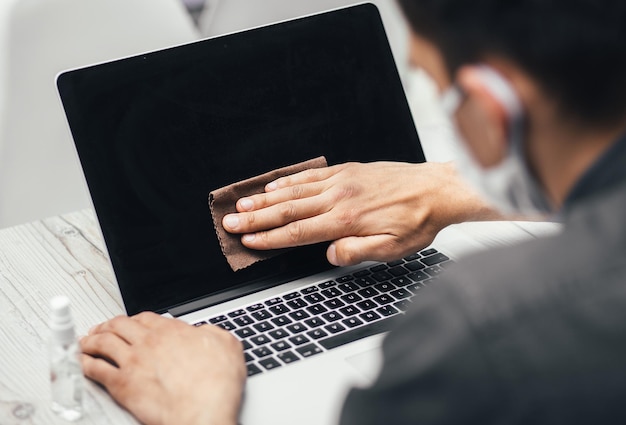 Close up. a young man in a protective mask wiping the screen of his laptop. personal hygiene and health care