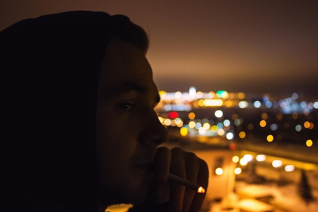 Photo close-up of young man smoking cigarette against illuminated city at night