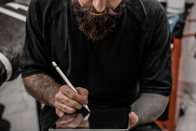 Photo close-up young man tattoo artist with beard drawing pencil and sketch standing in workshop place.