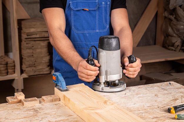 close up of the an young man wearing safety goggles carpenter builder preworks the edges of the wooden board with a milling machine in the workshop wooden sawdust flies to the sides