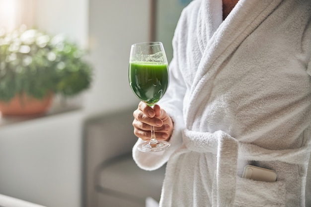 Photo close up of young man with healthy vitamin drink in his hand wearing white soft bathrobe