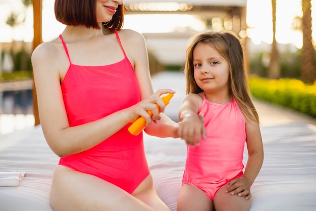 Close up of young mom in a pink swimsuit spraying SPF 50 sun protection cream on her daughter hand