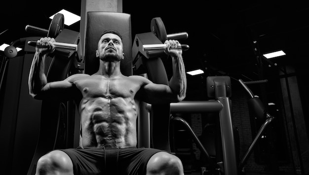 Close up of young muscular man sitting on simulator in gym.