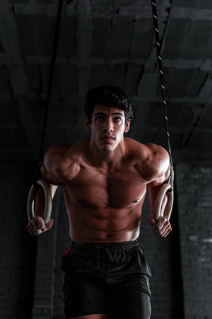 Close up of a young shirtless muscular athlete doing ring dips at a gym