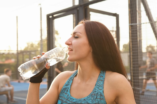 Close up of a young sportswoman drinking water on street workout playground on sunset