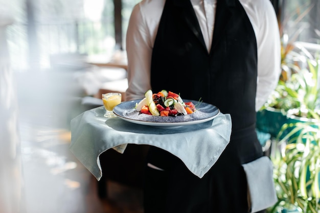 Close-up of a young waiter in a stylish uniform carrying an exquisite salad to a client in a beautiful gourmet restaurant. Table service in the restaurant.