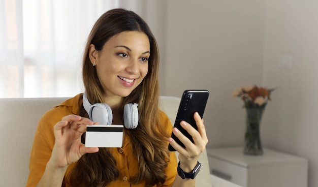 Close up of young woman holding credit card and using smart phone at home. Online shopping, spending money, enjoying life concepts.