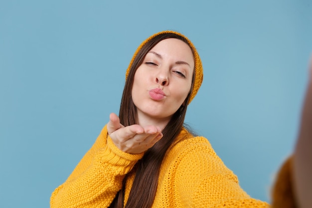 Close up of young woman in sweater and hat posing isolated on blue background studio portrait. People lifestyle concept. Mock up copy space. Doing selfie shot on mobile phone blowing sending air kiss.