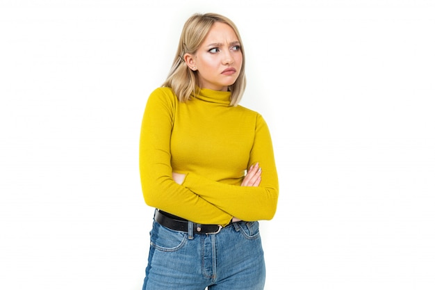 Photo close-up of a young woman with blonde hairstyle dressed in jeans and a yellow sweater with an indignant look on her face on a white isolated background