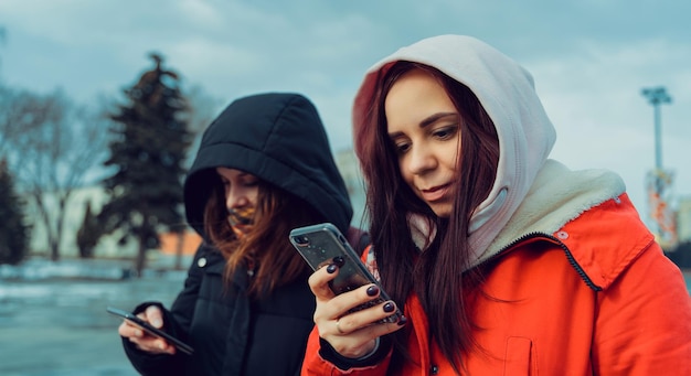 Close up of young women browsing smartphone in city park Pretty females in hoods using mobile phone