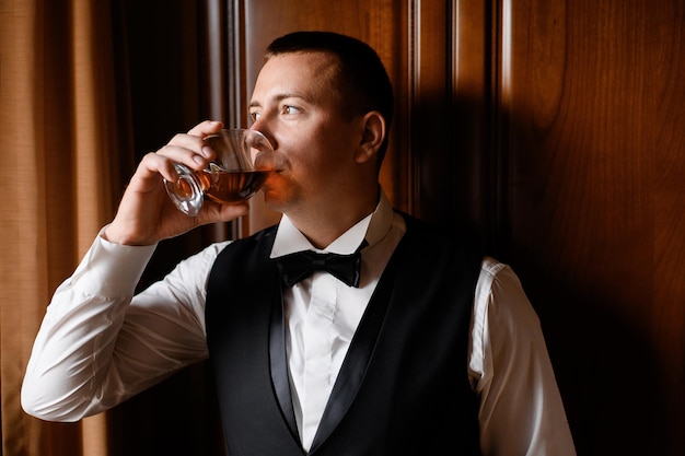 Close view of confident and handsome groom in trendy suit standing near vintage furniture seriously looking away while drinking alcohol in apartment during wedding morning