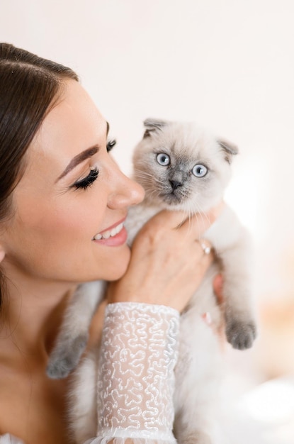 Close view of happy girl with natural makeup and hairdo wearing in elegant white dress smiling and looking to purebred Scottish fold kitty which she holding near her face