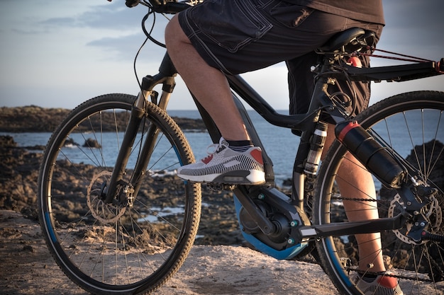 Close view of the legs of a senior man standing on the cliff with the electric bicycle. Healthy sport and lifestyle. Blue ocean water on background.