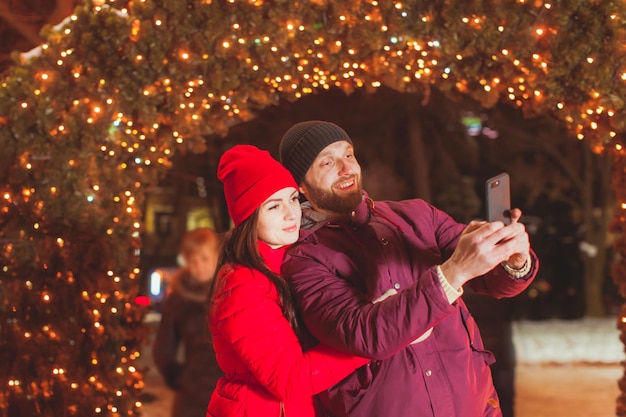 Close view portrait of young couple making selfie near fir tree arch, decorated with garlands. Married couple photographing while winter holiday traveling