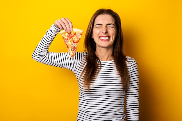 Closed eyes young woman smiling with closed eyes holding a slice of hot fresh pizza on a yellow background