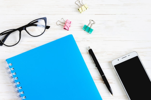 Closed notebook, pen, glasses, mobile phone, a cup of coffee on a white wooden table, flat lay, top view. Office table desk, workplace