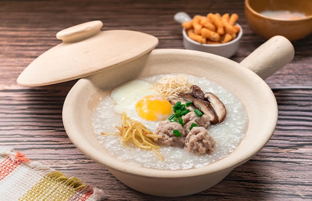 Closed up rice congee topped soft boiled egg boiled pork patty balls ginger and shiitake mushroom on white clay pot side dish by crispy deep fried dough stick