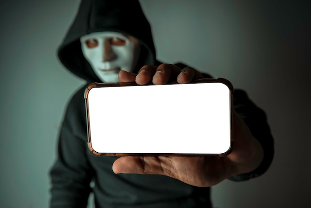 CloseUp An anonymous hacker wearing a mask and a black hoodie shows off a smartphone with a blank white screen Hacking and Malware Concepts