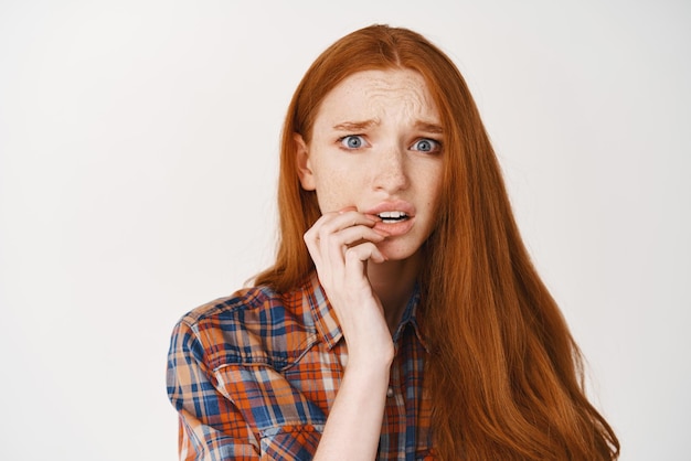 Closeup of anxious young woman with ginger hair biting finger and staring nervous at camera having worries standing over white background