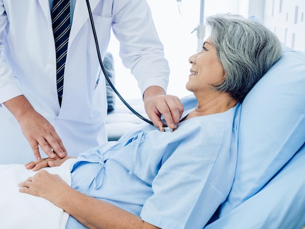 Closeup Asian elderly female patient dressed in light blue smiling happily in bed while the kindly male doctor in white suit uses stethoscope to listen to heartbeat for checkup in hospital