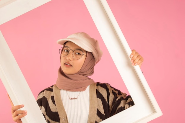 closeup of asian teen woman posing with white frame and wearing hijab, white hat and glasses.