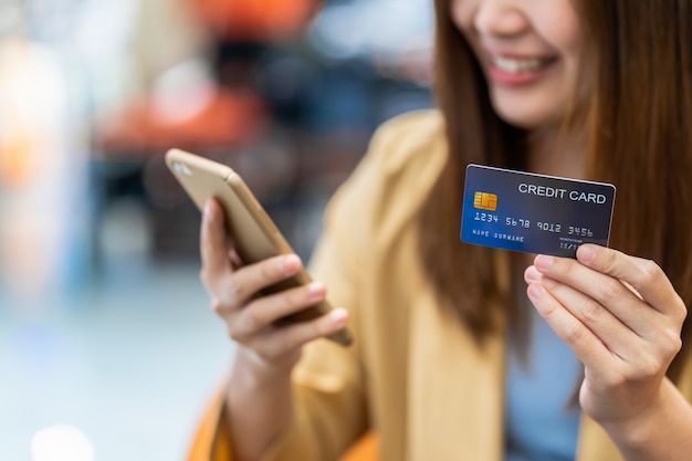 Closeup Asian woman hand holding the credit card and presenting the mobile phone for online shopping over the clothes shop store wall, technology money wallet and online payment concept