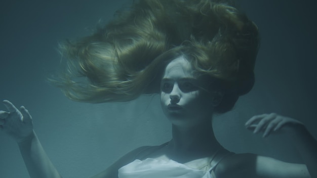 Closeup of a beautiful girl with red hair in a white dress froze under water