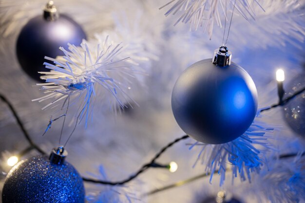 Closeup of beautiful shiny blue holiday baubles hanging on a white christmas tree with lights.