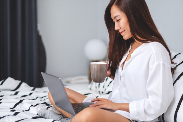 Closeup  of a beautiful woman using and working on laptop computer , drinking coffee while sitting on a white cozy bed at home