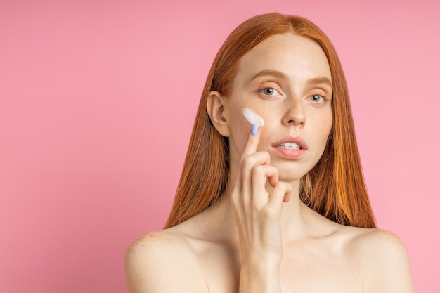 Closeup beauty portrait of redhead caucasian beautiful female applying moisturizing cream or face primer and looking at camera over pink background. Youth and skin care concept.