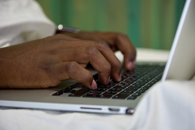 Closeup of a black African man typing with his hands on a laptop computer sitting on a white bed