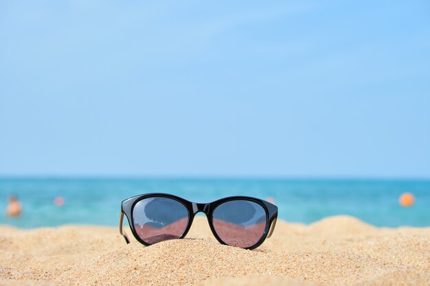 Closeup of black protective sunglasses on sandy beach at tropical seaside on warm sunny day. Summer vacation concept.