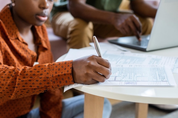 Closeup of blackwoman in orange shirt filling tax papers at coffee table