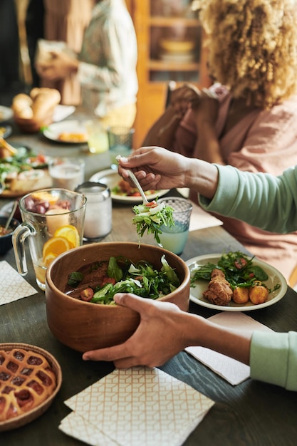 Closeup of boy putting fresh vegetable salad from bowl on his plate during dinner at table with his