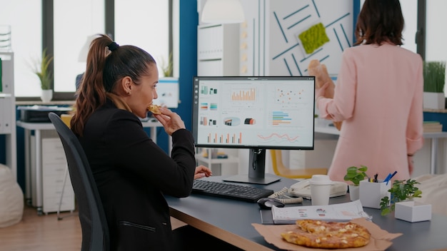 Closeup of businesswoman eating tasty pizza while analyzing financial statistics at desk in workplace