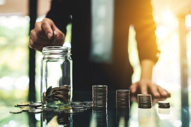 Closeup  of a businesswoman putting coins in a glass jar with coin stacks on the table for saving money and financial concept