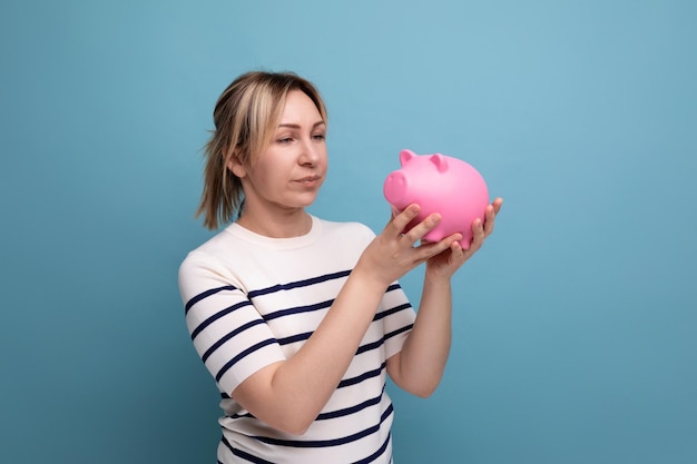 Closeup of a charming cute young woman in a casual outfit with a pink piggy bank of money on a blue