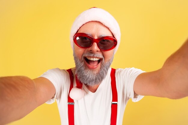 Photo closeup cheerful positive man in santa claus hat and stylish sunglasses looking into camera, posing making selfie. indoor studio shot isolated on yellow background