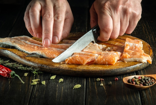 Closeup of a chef hands cutting fresh red salmon fish with a knife on a kitchen cutting board Cooking a delicious fish dish according to an old recipe with spices and pepper