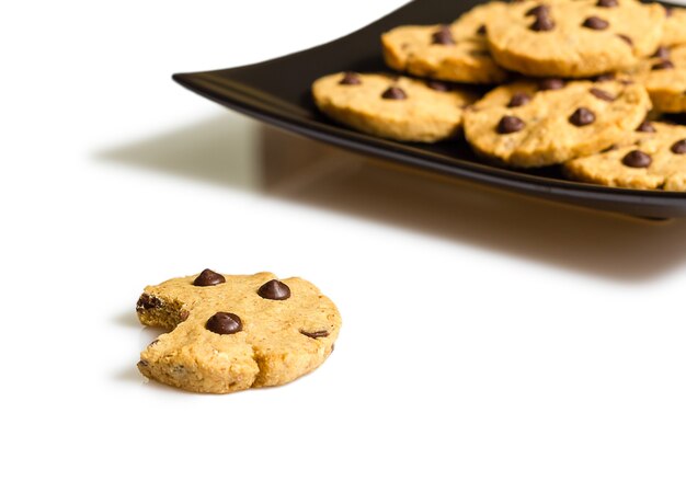 Closeup of chocolate chip cookie with a bite and pile of cookies in a square black plate on white background