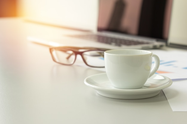 closeup coffee cup on table with laptop, glasses and papers in background